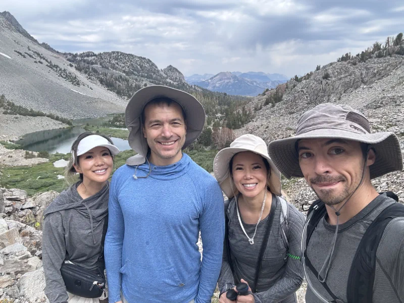 Group photo of four hikers at a dark alpine lake with granite mountains and snow patches
