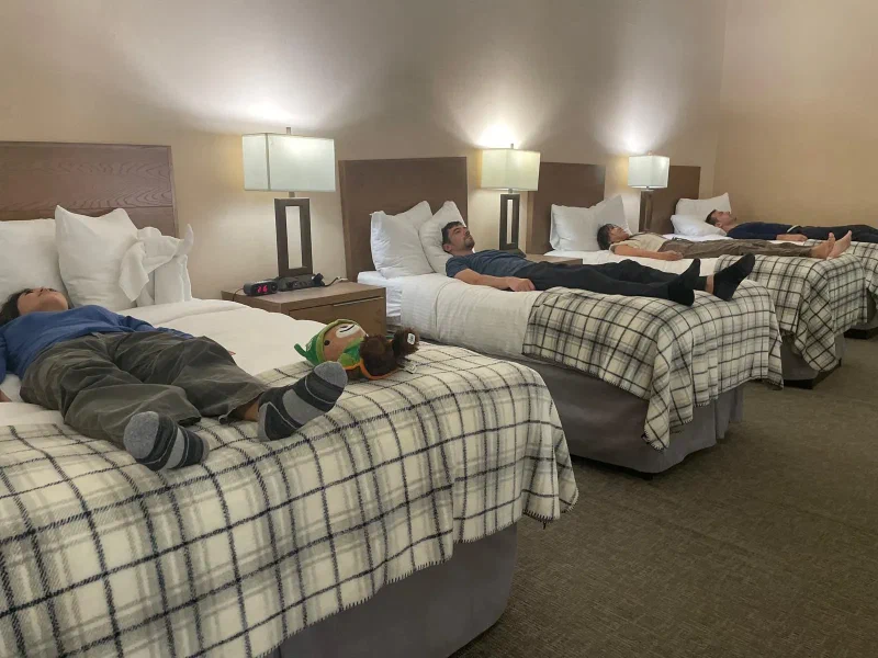 Five travelers lying flat on their backs across four twin beds in synchronized pose