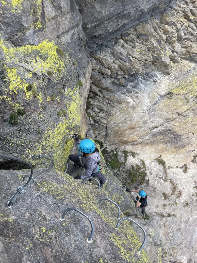 Two climbers in blue helmets ascending a via ferrata route on a vertical rock face