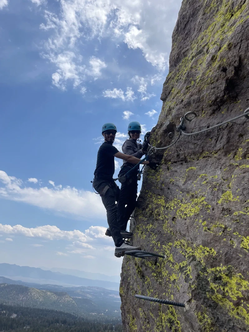Two climbers standing on metal rungs on a sheer cliff with a vast forested valley behind them