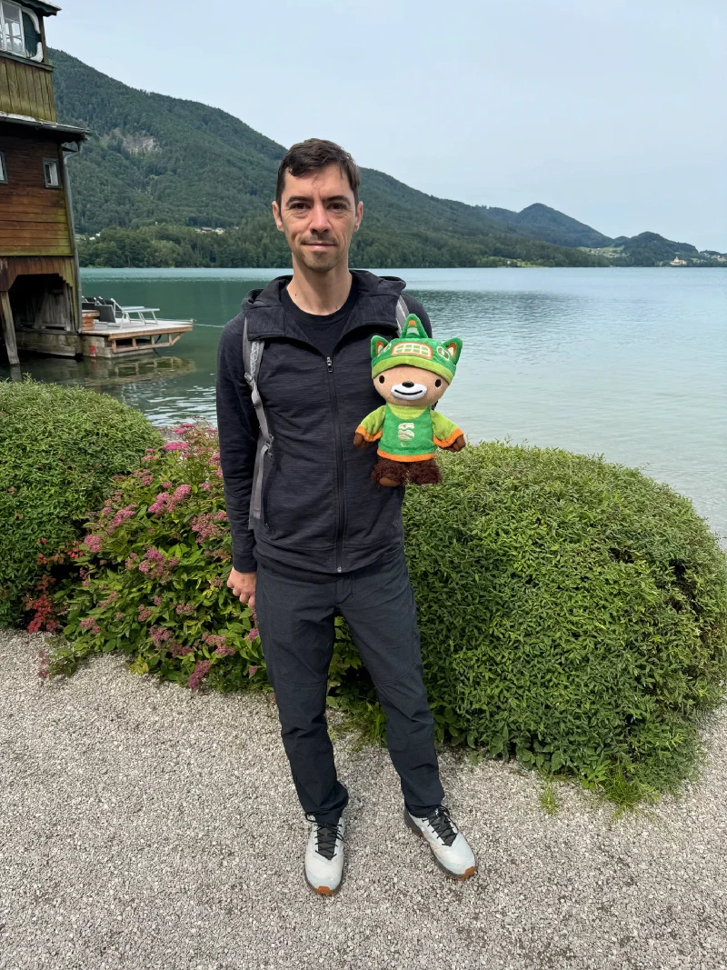 Nicholas holding Sumi by a calm alpine lake with forested mountains