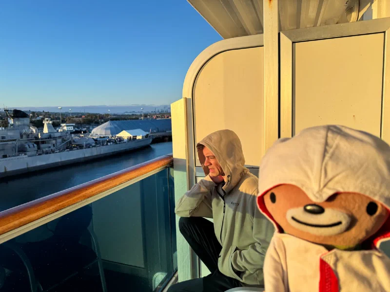 Nicholas crouched on the ship balcony at golden hour with Sumi, harbor and naval ship in background