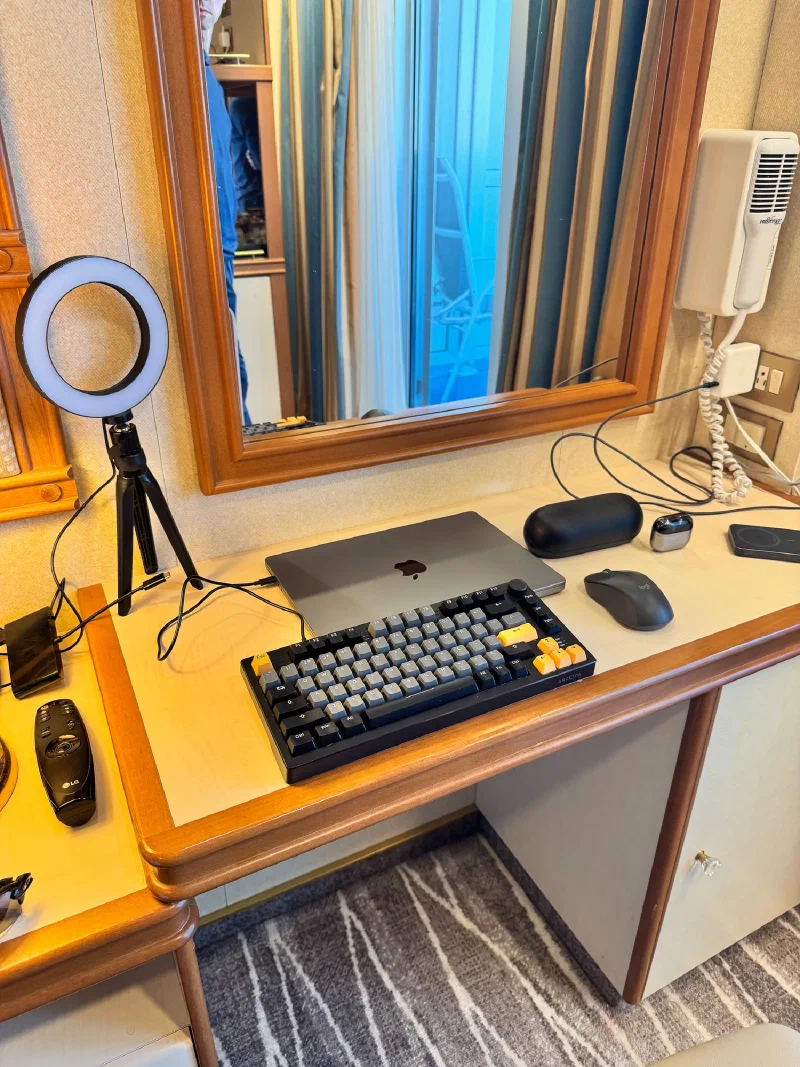 A cruise cabin desk completely taken over by laptop, mechanical keyboard, mouse, ring light, and chargers