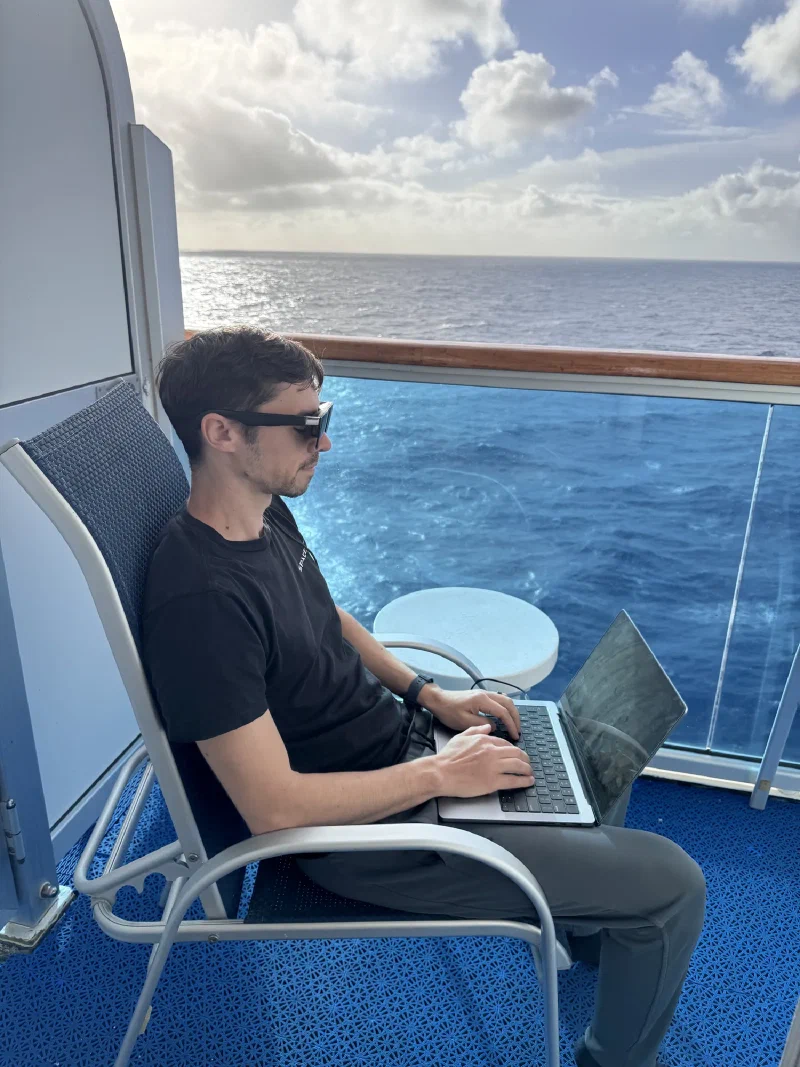 Nicholas sitting on cruise ship balcony with laptop, working with ocean view behind him
