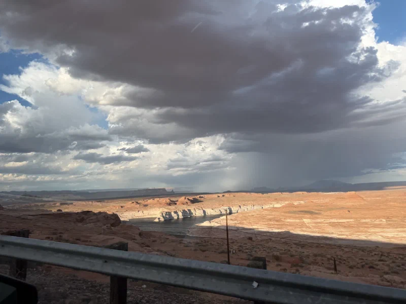 A dark storm cloud dropping a column of rain onto Lake Powell, with pale sandstone shores and red desert in the foreground, seen past a roadside guardrail