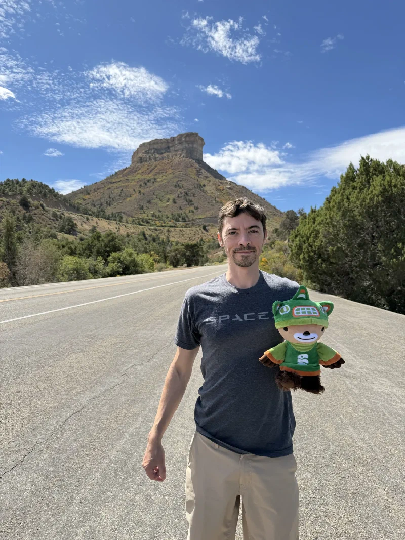 Nicholas in a grey SpaceX t-shirt holding Sumi on the shoulder of an empty road, with the flat topped Point Lookout mesa rising behind them