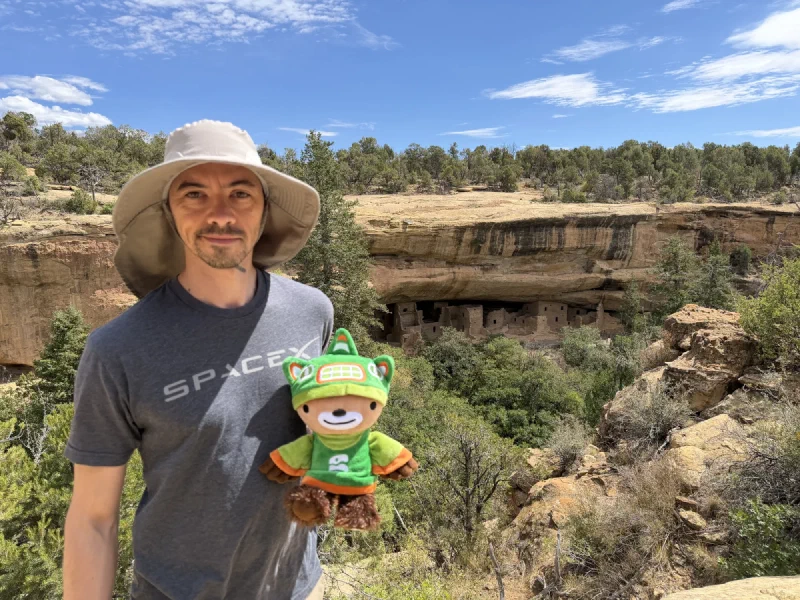 Nicholas in a wide brimmed sun hat with a neck flap and a SpaceX t-shirt holding Sumi, with the stone buildings of Spruce Tree House tucked under a sandstone cliff overhang in the canyon behind them