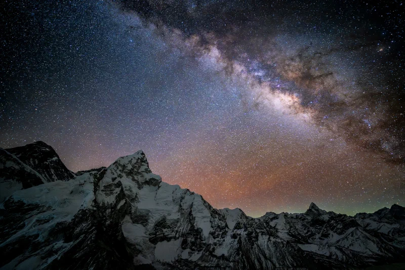 The Milky Way arcing over the jagged snow-covered peaks of the Everest massif from Kala Patthar, with orange and green airglow along the horizon