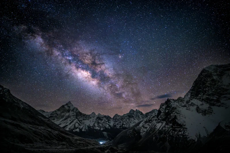 The Milky Way galactic core over the snow-capped peaks and dark valley of the Sherpa Memorial near Lobuche, with the distant lights of Dingboche on the valley floor