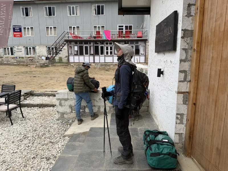 Nicholas standing on lodge steps with his pack, looking weary, under a sign reading The Himalayan Tyangboche