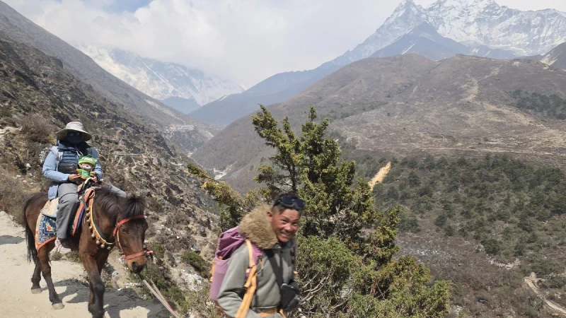 Po On riding a horse on a mountain trail with a guide walking alongside and snowy peaks in the distance