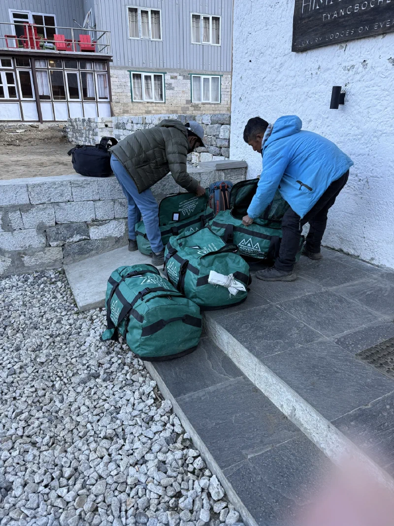 Two porters sorting through green duffel bags on a stone patio at a Tengboche lodge