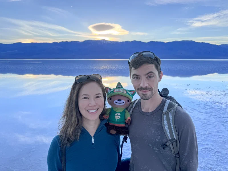 Nicholas and Pokin holding Sumi Bear at Badwater Basin at sunset with water reflecting mountains and lenticular clouds
