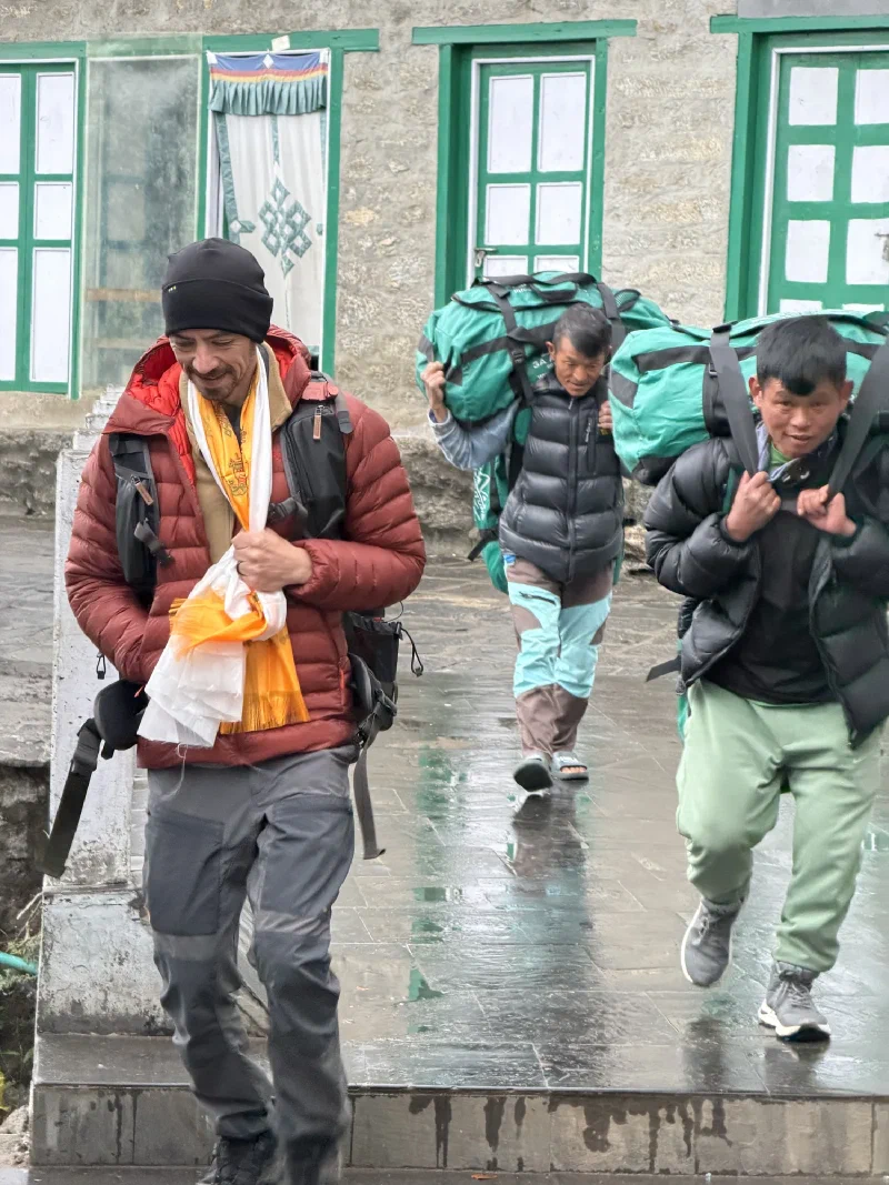 Nicholas and porters walking on a wet path in the rain toward the airport