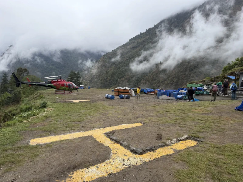 A grassy helipad with a yellow H, one red helicopter parked, low clouds on every mountain