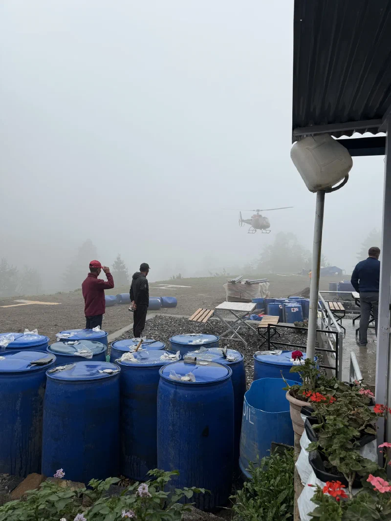 A Fishtail helicopter descending into extremely heavy fog over the barrel-strewn helipad, men watching