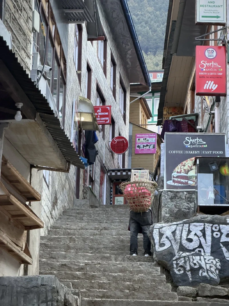Porter carrying heavy doko basket up steep stone stairs past Sherpa Barista and Baskin Robbins signs