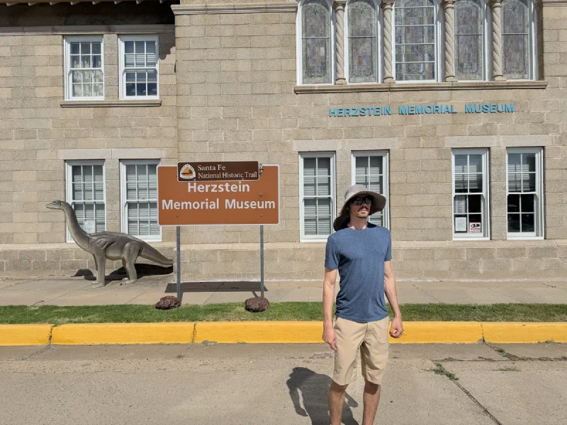 Nicholas in a bucket hat standing outside the Herzstein Memorial Museum in Clayton, New Mexico, beside a life-sized dinosaur statue