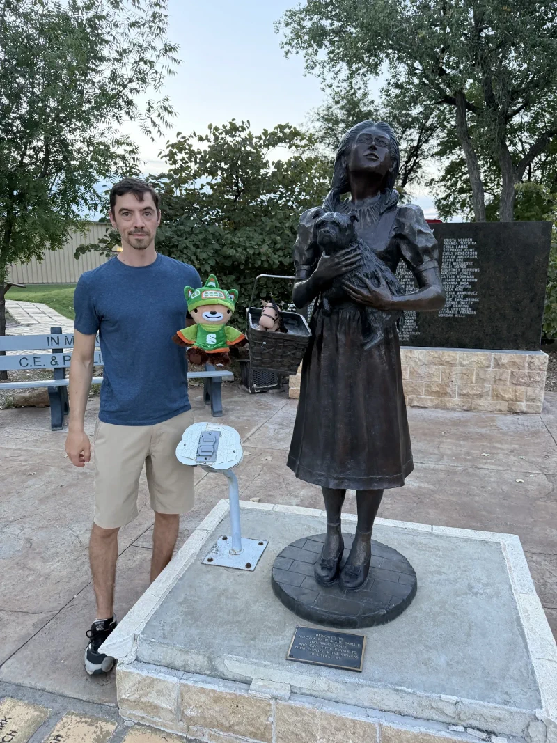 A bronze statue of Dorothy holding Toto on a stone base with a dedication plaque, in a park setting