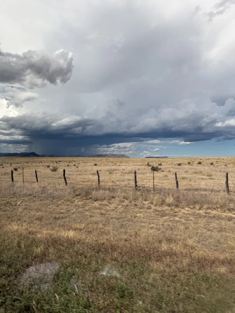 A wire fence and dry grass field stretching to the horizon under an enormous dark storm cloud with a rain shaft falling in the distance