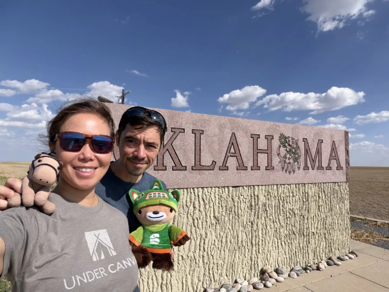 Pokin and Nicholas in a selfie at the Oklahoma state line sign, holding Sumi and Chestnut the toy horse up to the camera