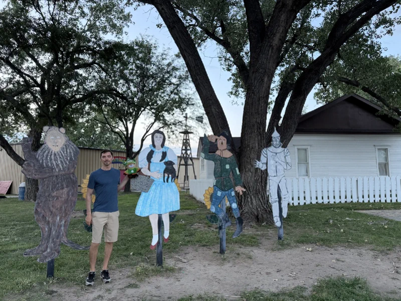 Nicholas holding Sumi beside four painted plywood cutouts of the Cowardly Lion, Dorothy, the Scarecrow and the Tin Man, with a white farmhouse and windmill behind