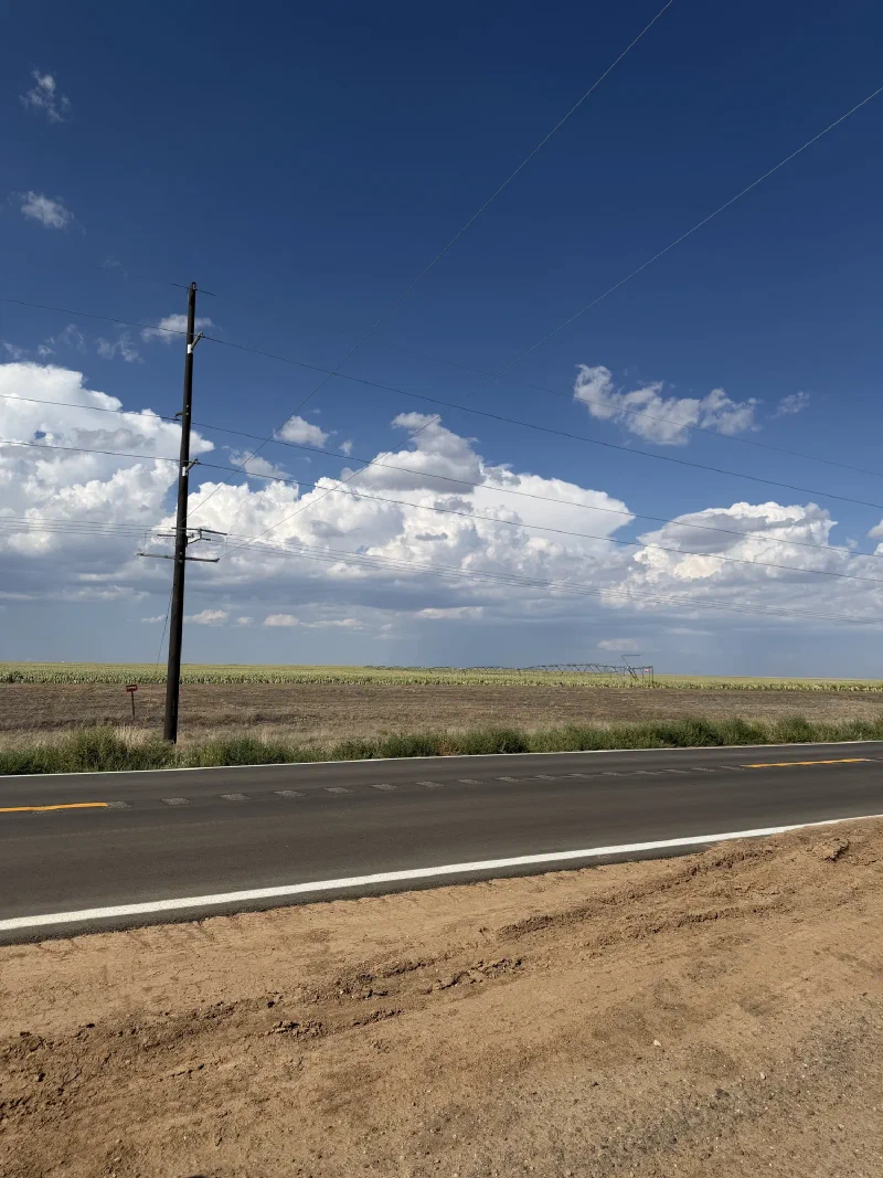A two-lane road with a yellow centreline running past green irrigated fields under a deep blue sky with scattered cloud