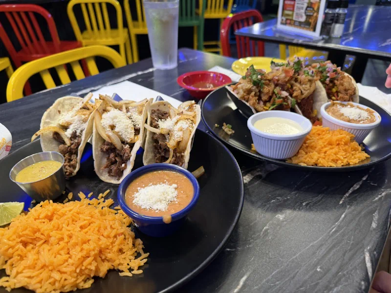 Two plates of tacos with rice and small bowls of salsa on a table, with bright yellow red green and blue chairs in the background
