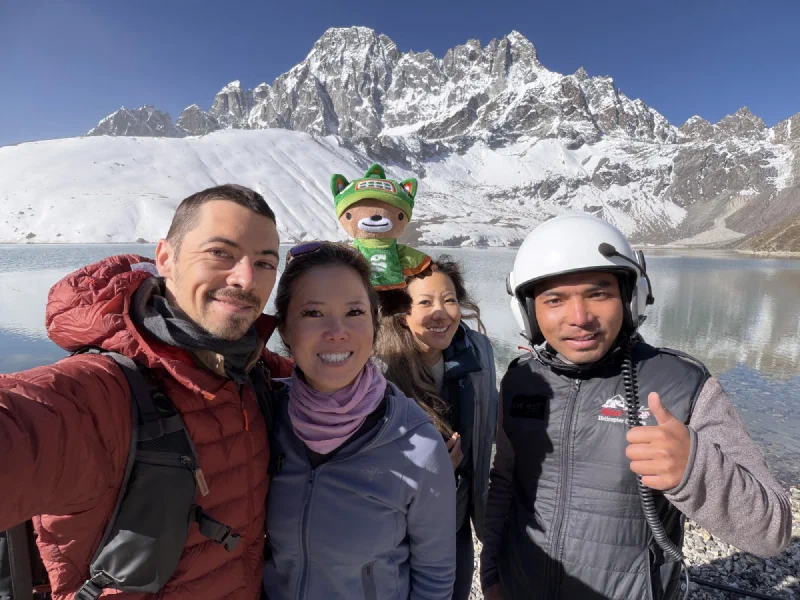 Group selfie with helicopter pilot giving thumbs up at Gokyo Lake, snowy mountains behind