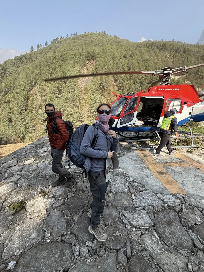 Two trekkers stepping away from a red helicopter on a rocky helipad with forested hillside behind