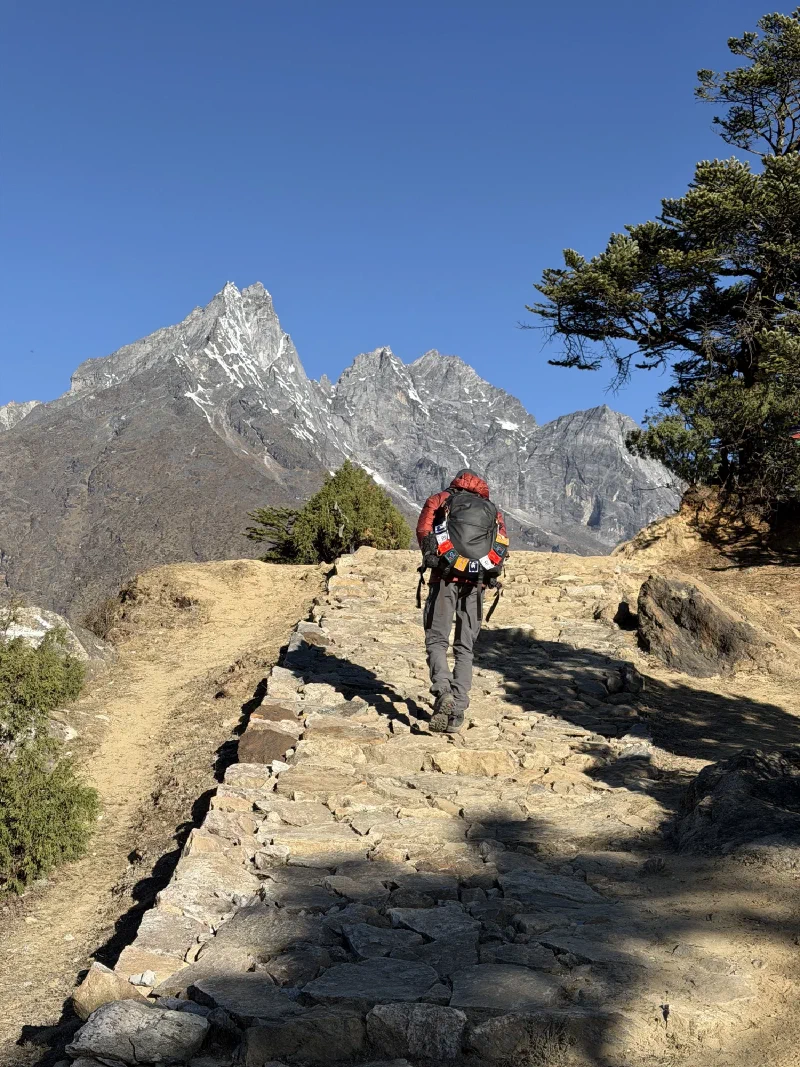Nicholas hiking uphill on a rocky trail with a red jacket and large backpack, mountains behind