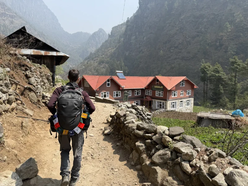 A trekker walking down a stone path toward a lodge with red roofs in a dramatic valley setting