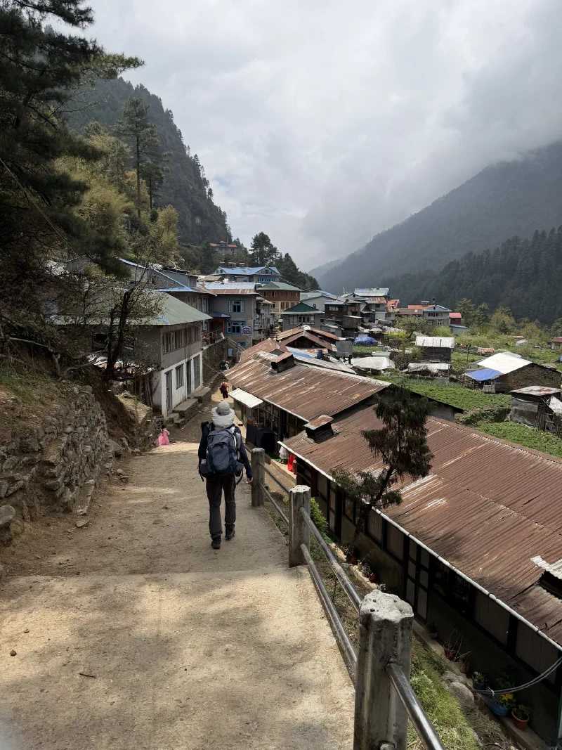 A trekker descending a dusty village path with mountains in the cloud behind