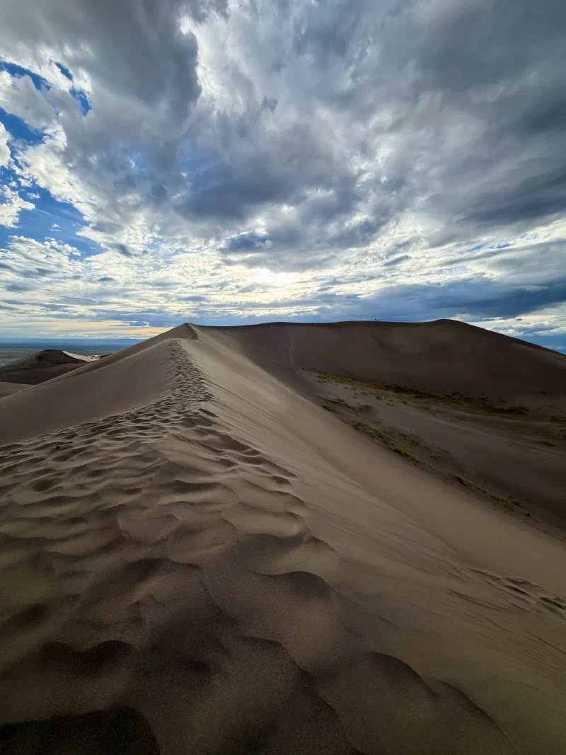 A knife edge dune ridge covered in footprints leading up to a crest, with a further higher crest beyond it under heavy cloud