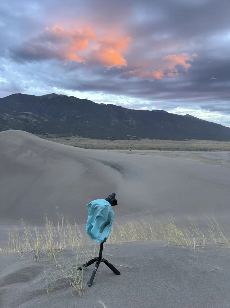 A camera on a tripod wearing a teal rain cover on the dunes at dusk with pink lit cloud over the mountains