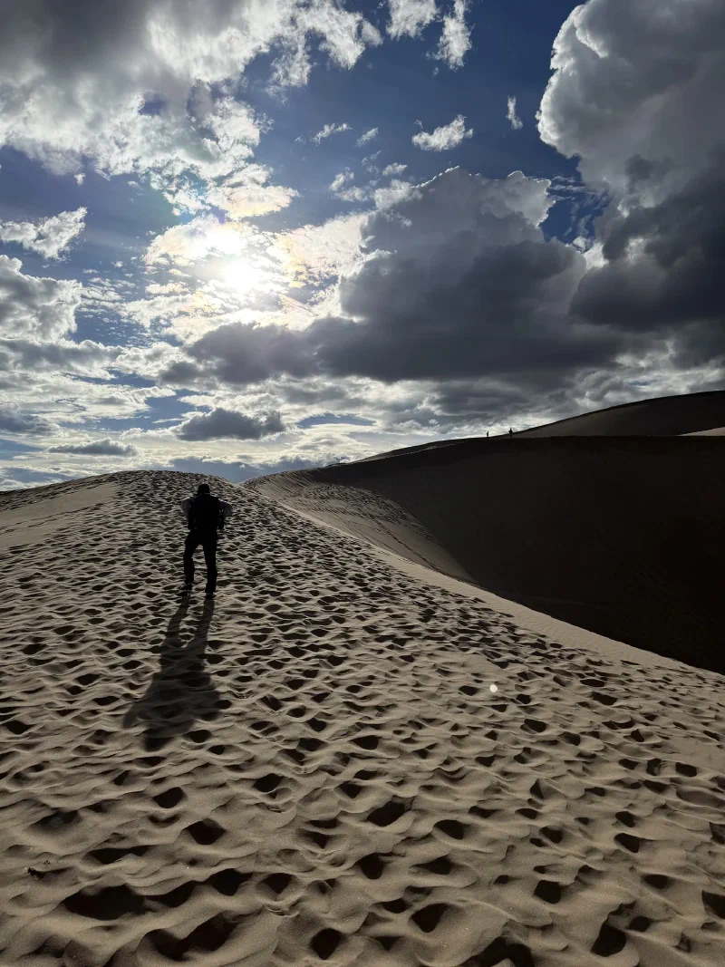 Nicholas silhouetted climbing a sharp dune ridge with the sun breaking through a dramatic bank of storm cloud above him