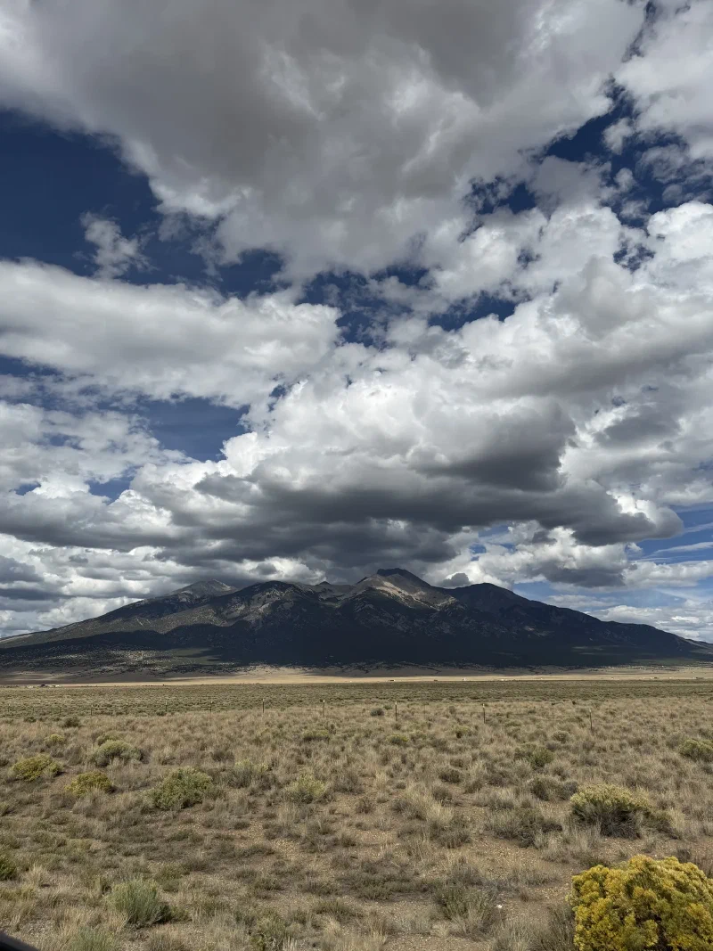 A dark mountain massif rising from a flat sagebrush valley under an enormous bank of grey and white cloud