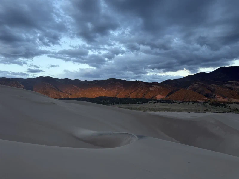 The dunes in shadow with the Sangre de Cristo mountains catching the last orange light under a heavy grey sky