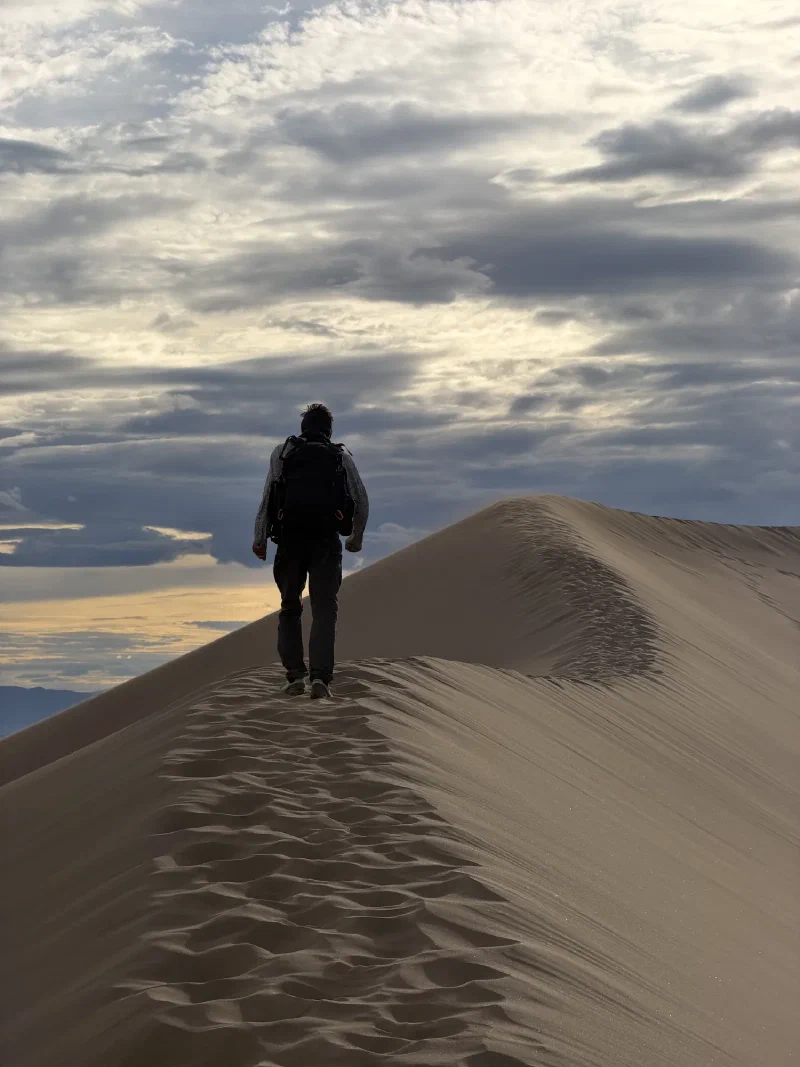 Nicholas walking away along the crest of a dune with a backpack, silhouetted against soft evening cloud