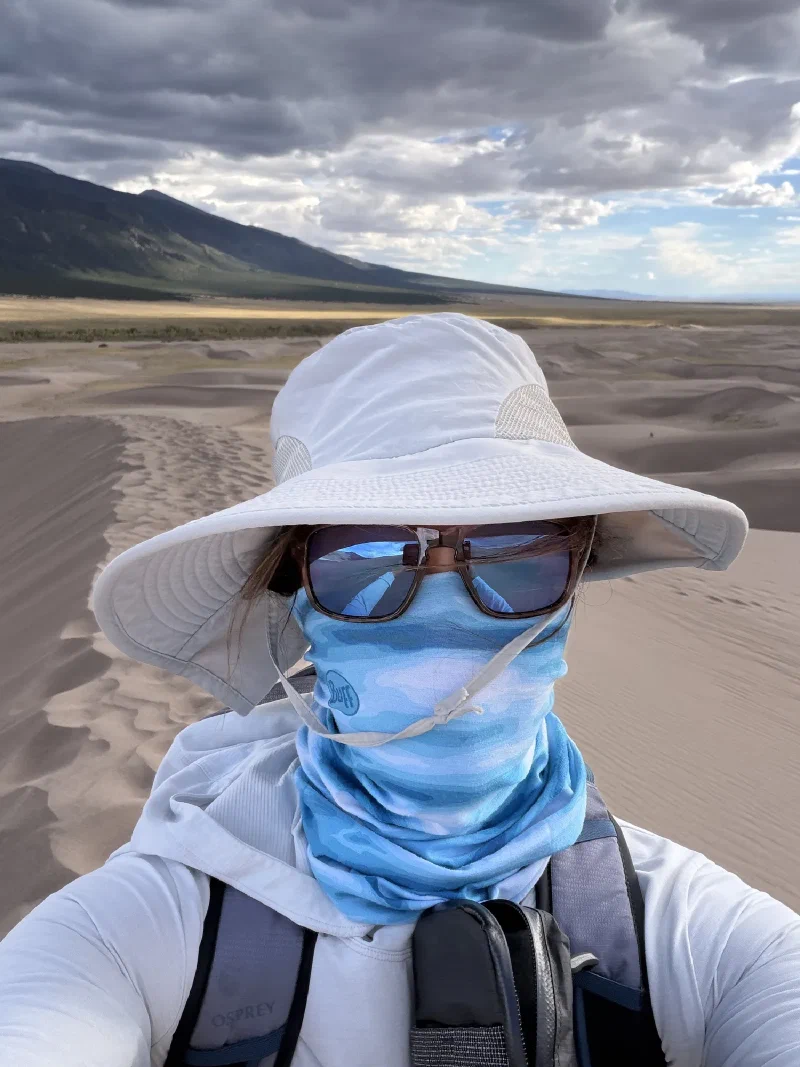 Pokin in a wide white sun hat, sunglasses and a blue buff pulled over her face, dunes and dark cloud behind her