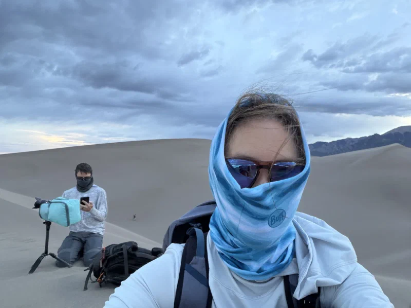 Pokin with her buff pulled right up over her nose and head, eyes barely visible behind sunglasses, while Nicholas kneels at his tripod behind her
