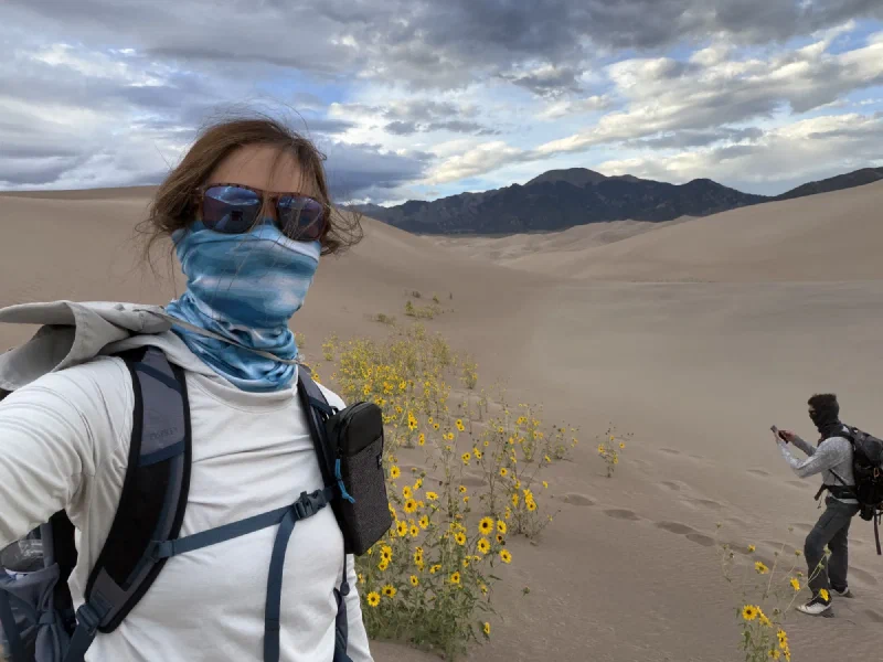 Pokin in her buff and sunglasses in the foreground with dune sunflowers and Nicholas photographing behind her, mountains in the distance