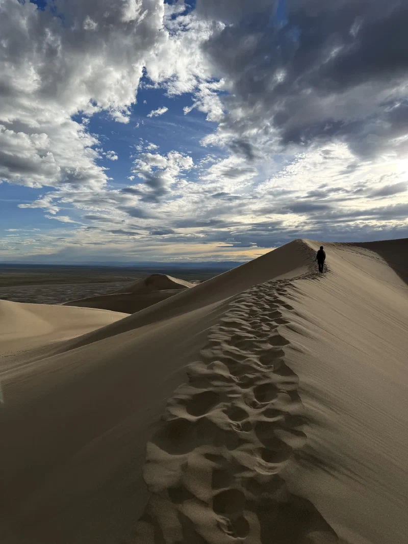 A long dune ridge covered in footprints leading to a tiny distant figure, with dunes and valley stretching out under a huge sky