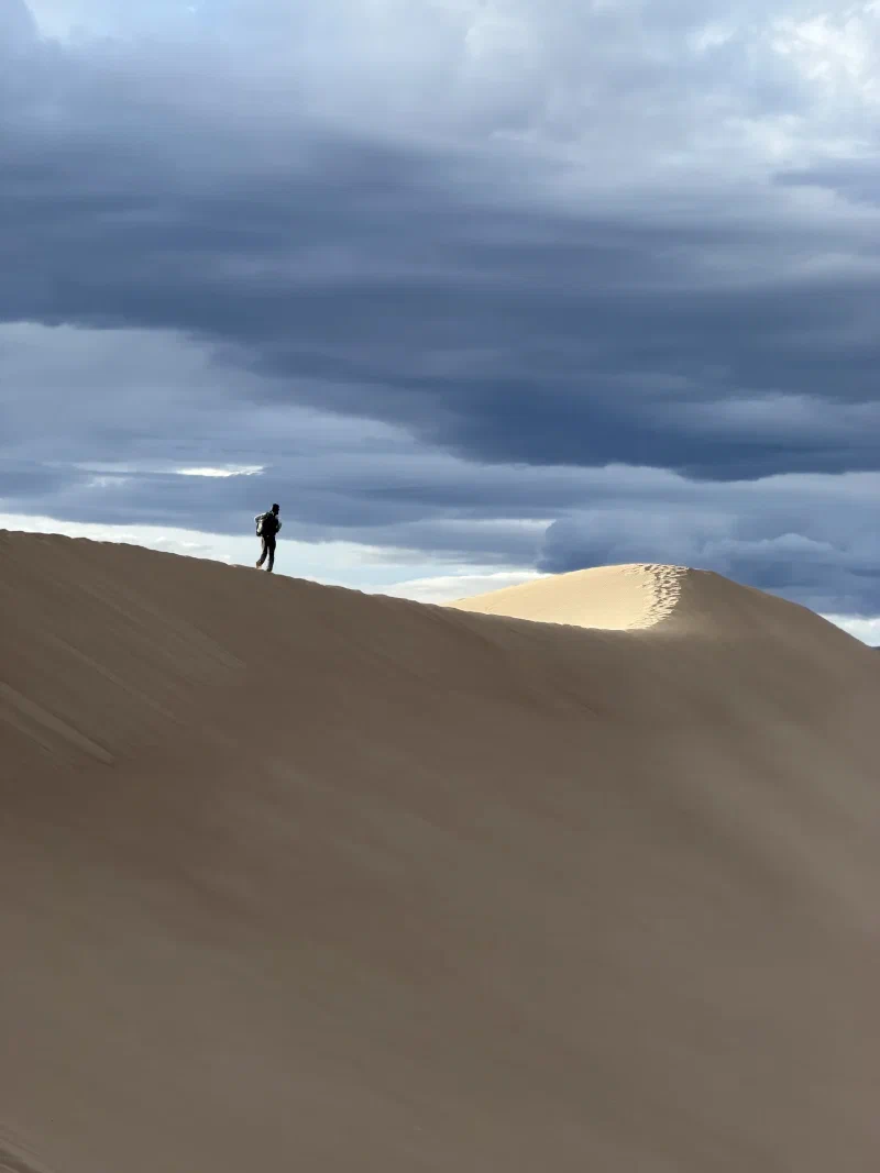 A lone figure on a vast dune ridge beneath an enormous dark shelf of storm cloud with one sunlit dune crest glowing behind
