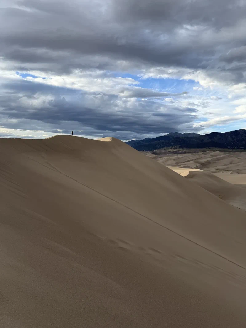 A tiny lone figure standing on a distant dune crest with the Sangre de Cristo mountains and heavy storm cloud behind