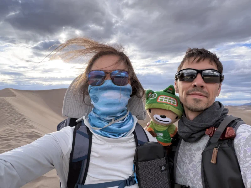 Pokin with her hair blowing sideways and her buff up, Sumi in the middle, and Nicholas in sunglasses, all on the dune ridge under grey cloud