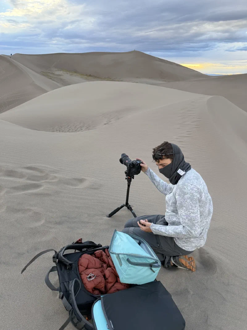 Nicholas kneeling beside a camera on a low tripod with gear bags open on the sand, his buff pulled up over his face