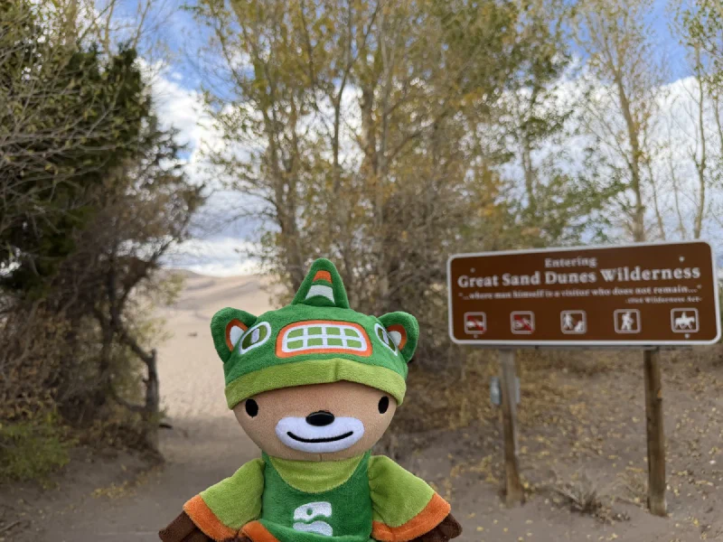 Sumi in his green toque and tunic propped in front of a brown sign reading Entering Great Sand Dunes Wilderness, with a dune rising behind the cottonwoods