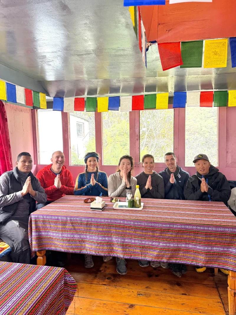 Full group Namaste photo at the lunch table with prayer flags on the ceiling