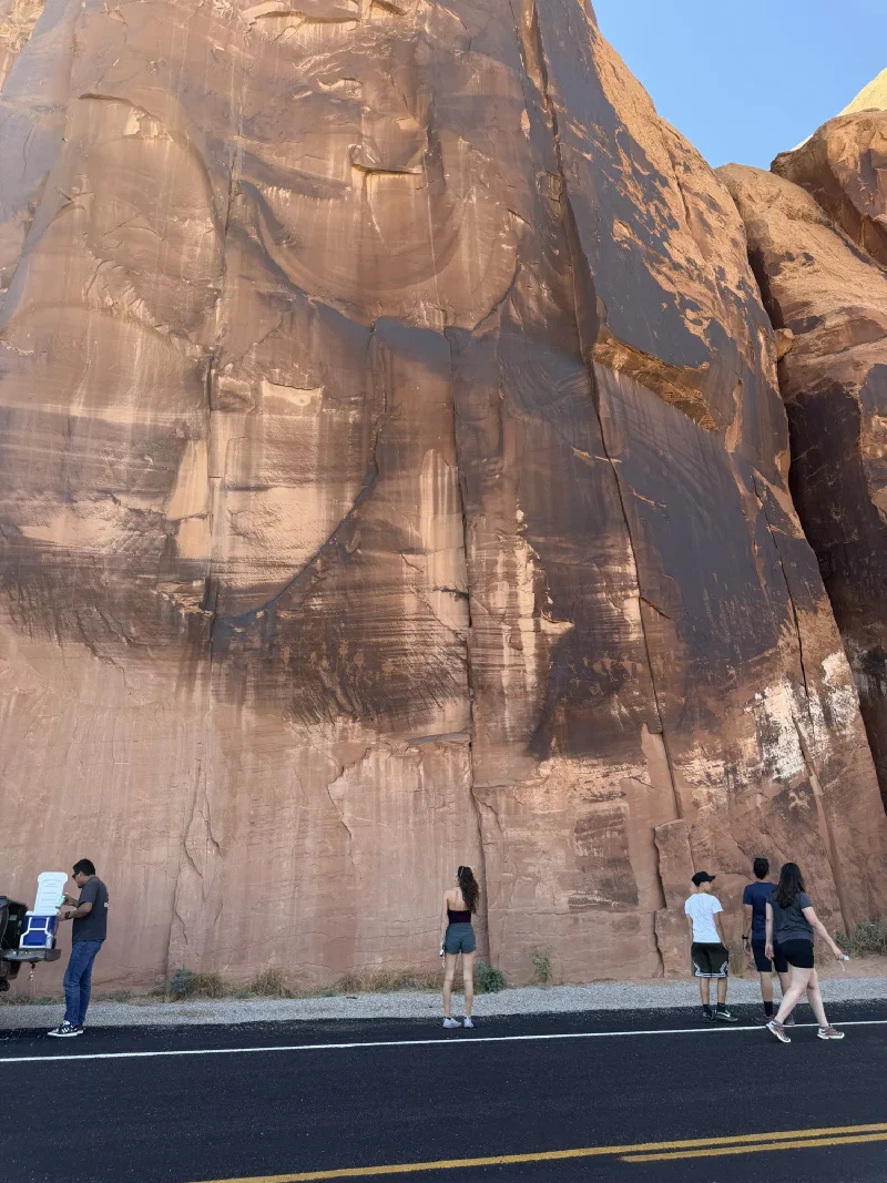 People standing at the base of a towering sandstone wall beside the road, looking up at petroglyphs